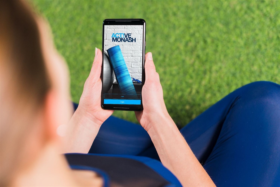 A woman in a gym attire holding a phone with the Active Monash on the screen.